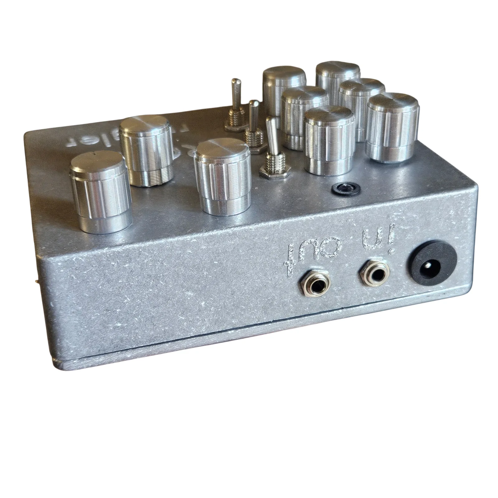 The box from its top edge at a low angle: two small jacks engraved in and out and a black DC socket along the edge, with the knobs and toggles standing up on the face behind them.