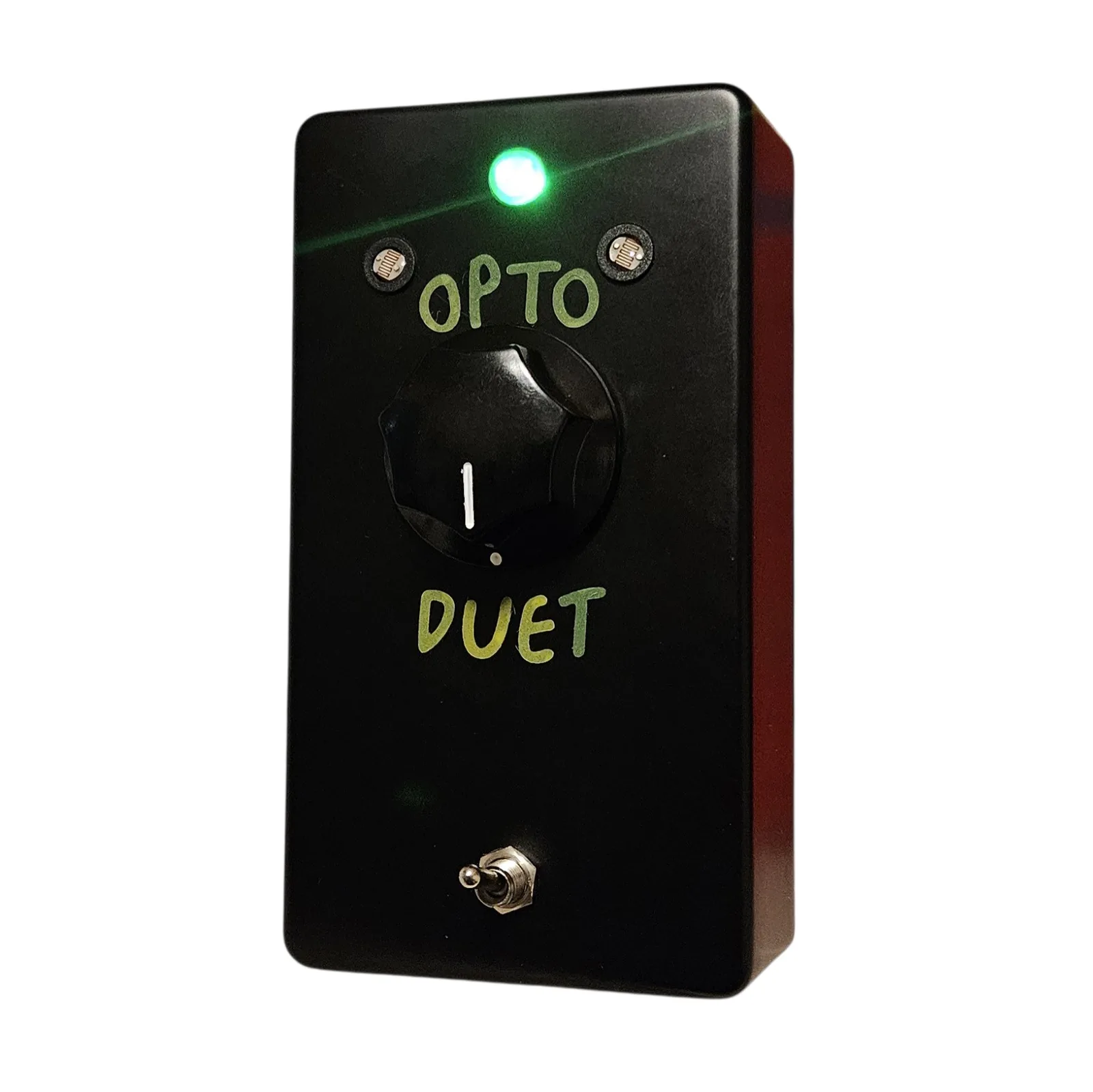 The same box with the toggle flicked up and the LED at the top lit, throwing a small halo across the face. The two round mesh-faced windows either side of it catch the light, and the OPTO DUET lettering and the fluted knob read clearly against the black.