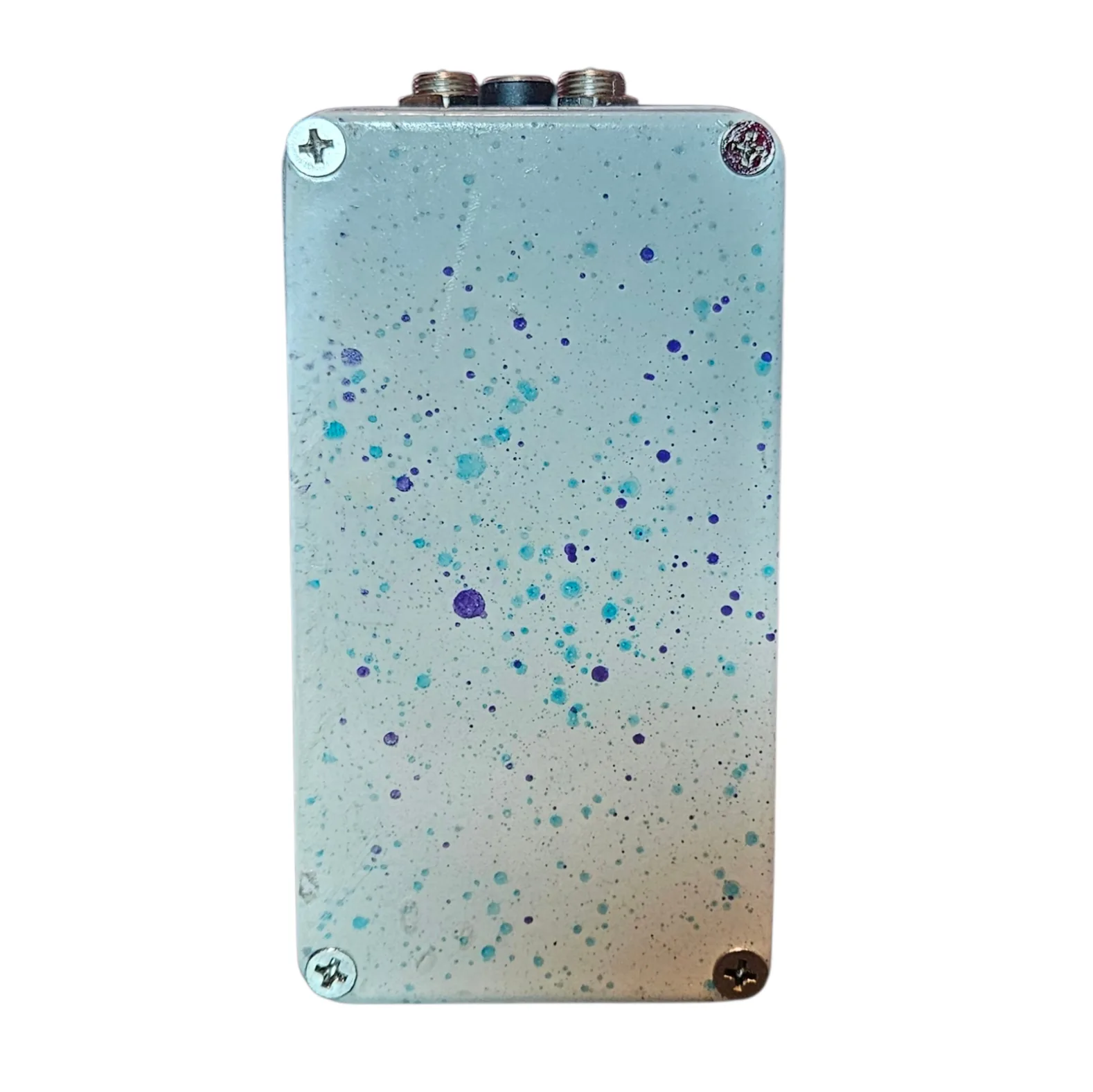 Back of the pedal: a cream bottom plate scattered with teal and purple splatter dots, with a Phillips screw in each corner and no label. Two jacks and the DC socket show along the top edge.
