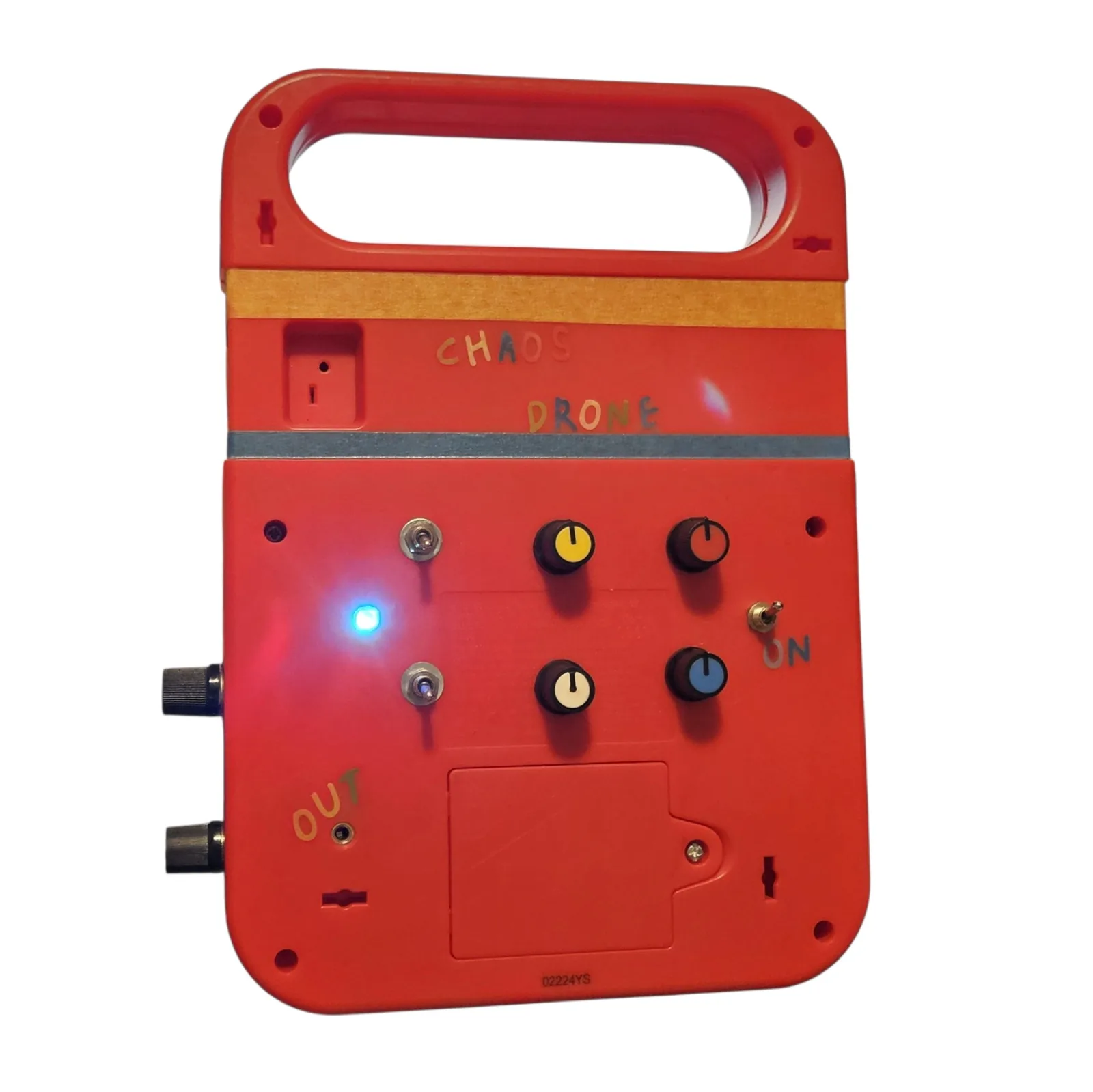 The back of the Speak & Spell, powered up. CHAOS and DRONE are spelled out in sticker letters across two strips of gold and gray tape. Below them are four black knobs with yellow, orange, cream and blue pointers, two silver toggle switches, a third toggle labeled ON in sticker letters, and a small blue indicator LED lit beside them. OUT is lettered next to a small jack at the bottom left, and two more knobs come through the left edge of the case.