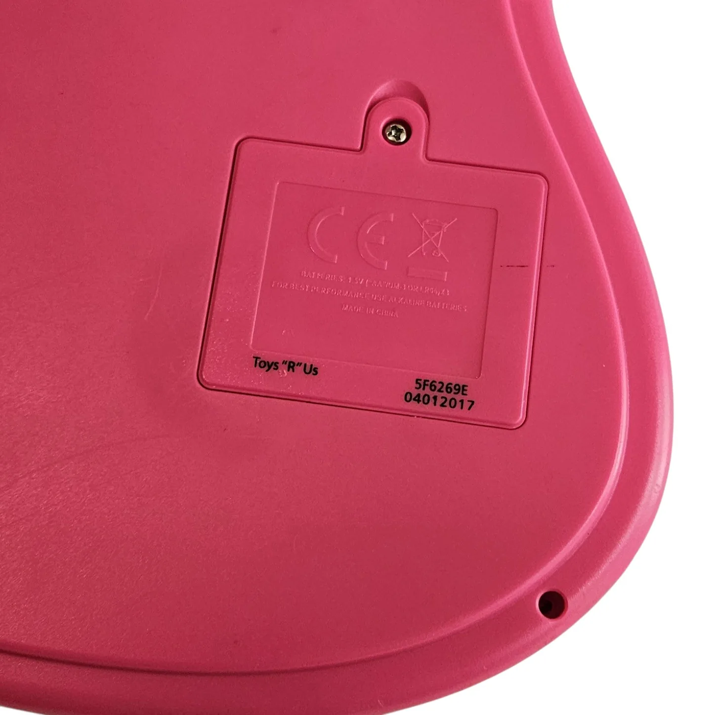 The lower back corner in close-up: pink molded plastic carrying a CE mark, small safety text and a Toys R Us model number, with a round hole near the bottom edge.