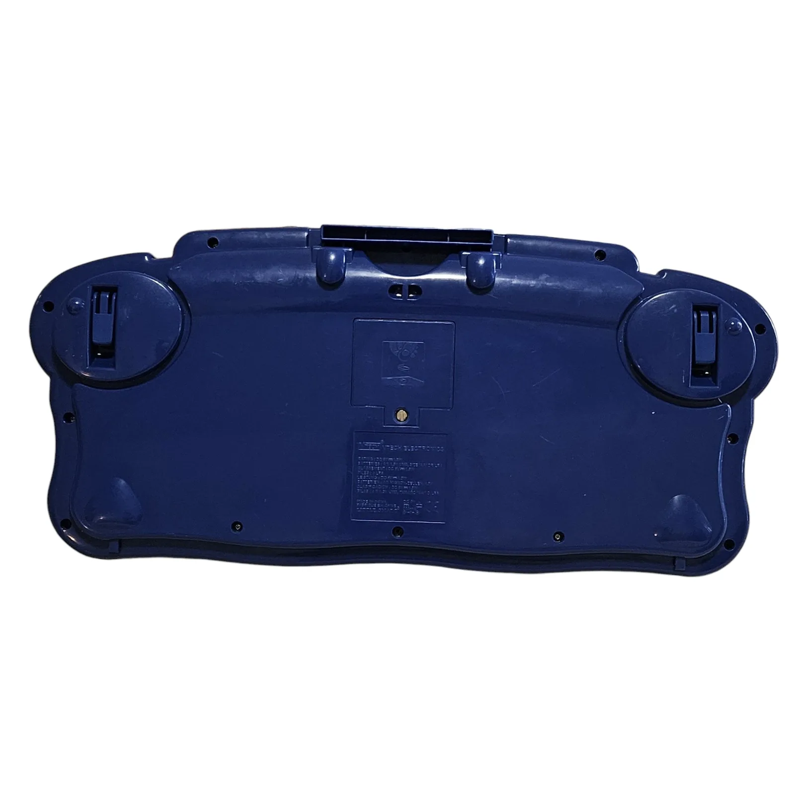 The underside, plain dark blue plastic with a battery door in the middle held by a single screw, VTech Electronics text and battery warnings molded into the plastic, the two round speaker housings at the outer corners, a fold-out stand along the top edge and four raised feet.