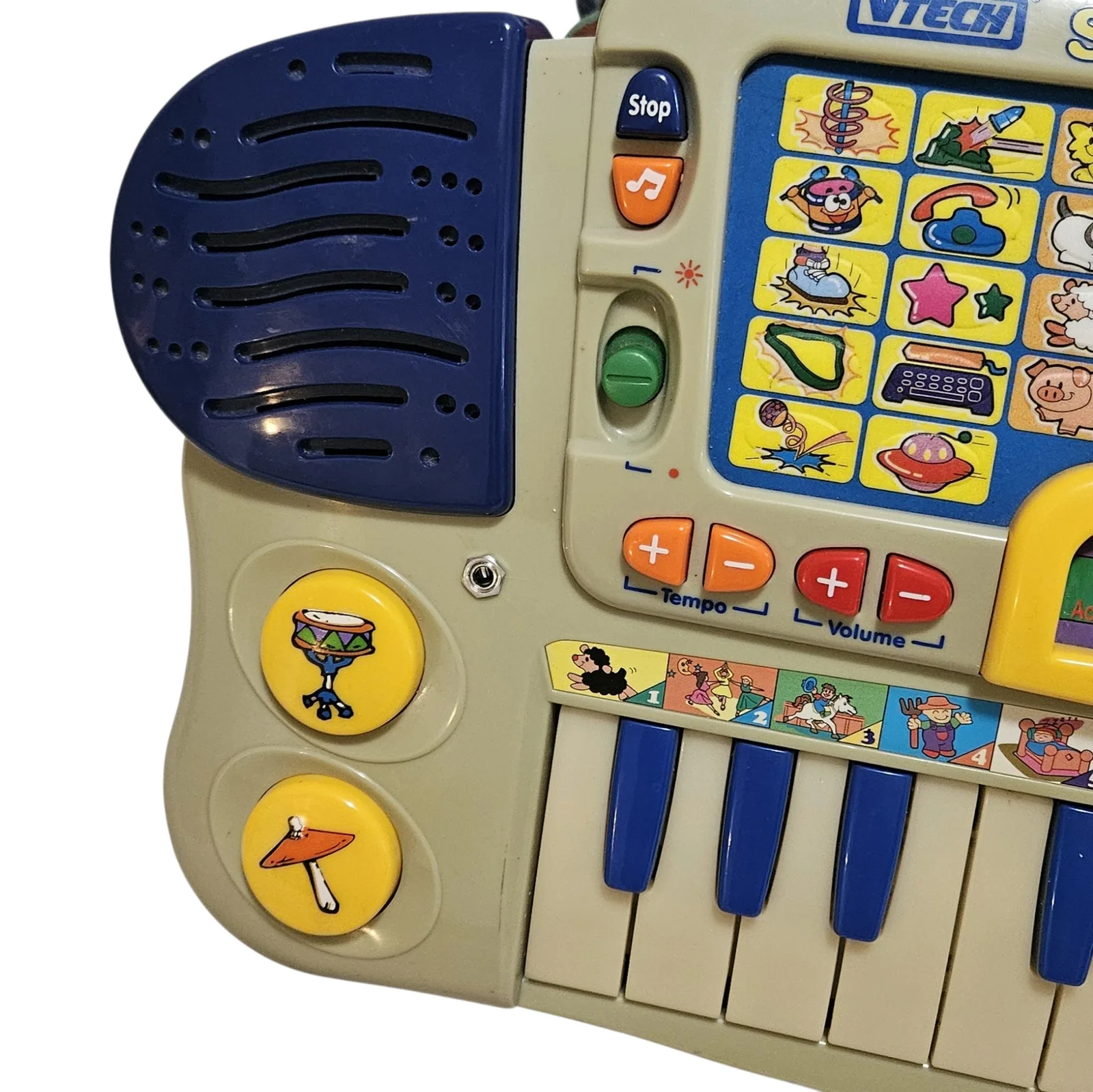 A close angled view of the left end. A small metal jack socket is set into the cream case between the two round yellow drum pads and the Tempo buttons: the added line out. Above it are the blue Stop button, the orange music-note button and a green slider, with orange Tempo and red Volume buttons to the right and the dark blue speaker grille above.