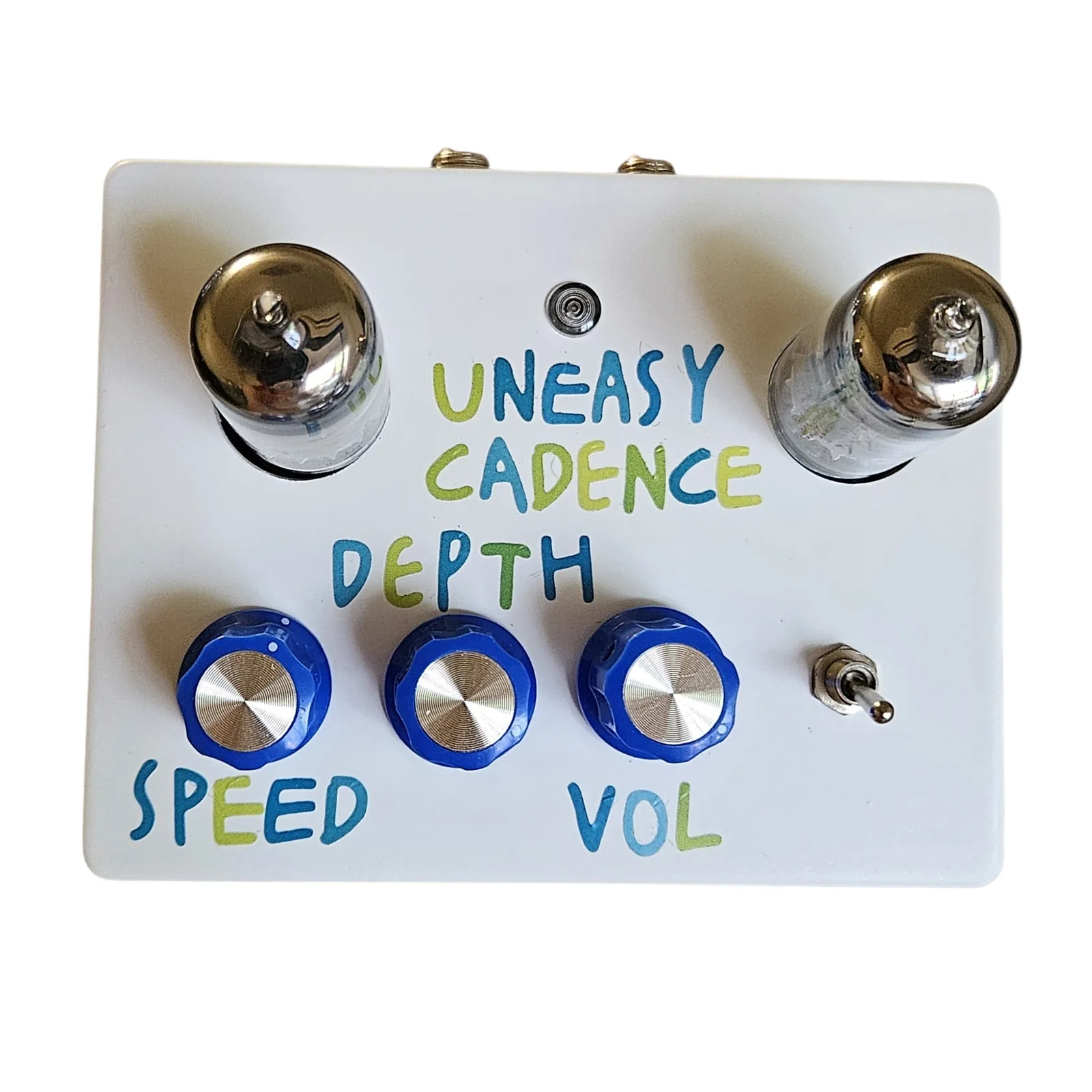 A white enclosure photographed face on from above. Two glass tubes stand up out of round holes at the top corners, with a small round indicator lamp between them and UNEASY CADENCE hand-lettered below it on two lines in blue and green. Three blue knobs with silver caps run along the bottom, labeled by hand: SPEED under the left knob, DEPTH above the middle one and VOL under the right. A small silver toggle sits to the right of the knobs, and the nuts of the two jacks show on the top edge.