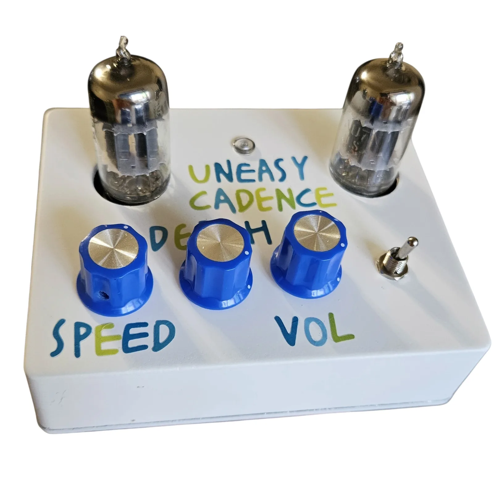 The face from in front at a low angle, the two tubes standing tall at the back corners with their metal insides visible through the glass. UNEASY CADENCE is lettered between them under the lamp, and the three blue knobs sit in a row in front with SPEED and VOL lettered below them and the toggle at the right.