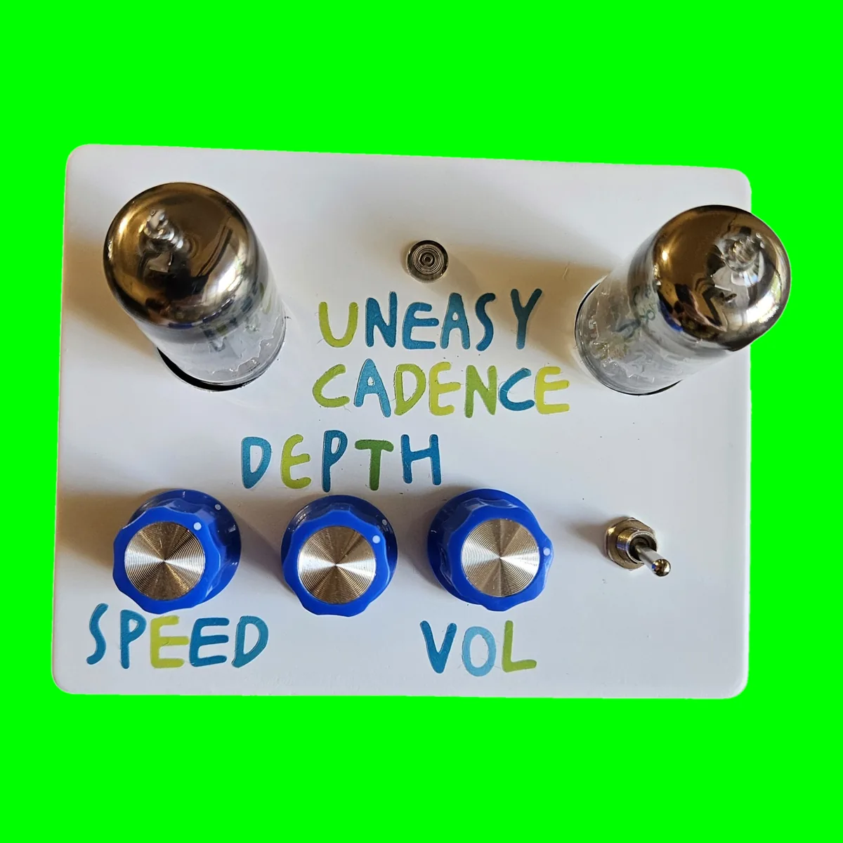 A white enclosure photographed face on from above. Two glass tubes stand up out of round holes at the top corners, with a small round indicator lamp between them and UNEASY CADENCE hand-lettered below it on two lines in blue and green. Three blue knobs with silver caps run along the bottom, labeled by hand: SPEED under the left knob, DEPTH above the middle one and VOL under the right. A small silver toggle sits to the right of the knobs.