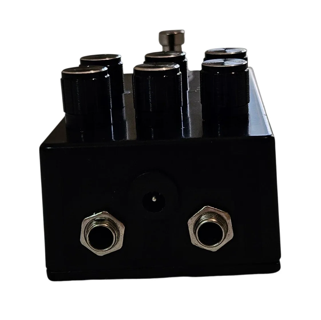 The top edge of the pedal straight on, with a silver quarter-inch jack socket at each end and a recessed barrel power socket centered between them. The six black knobs stand along the top of the frame.