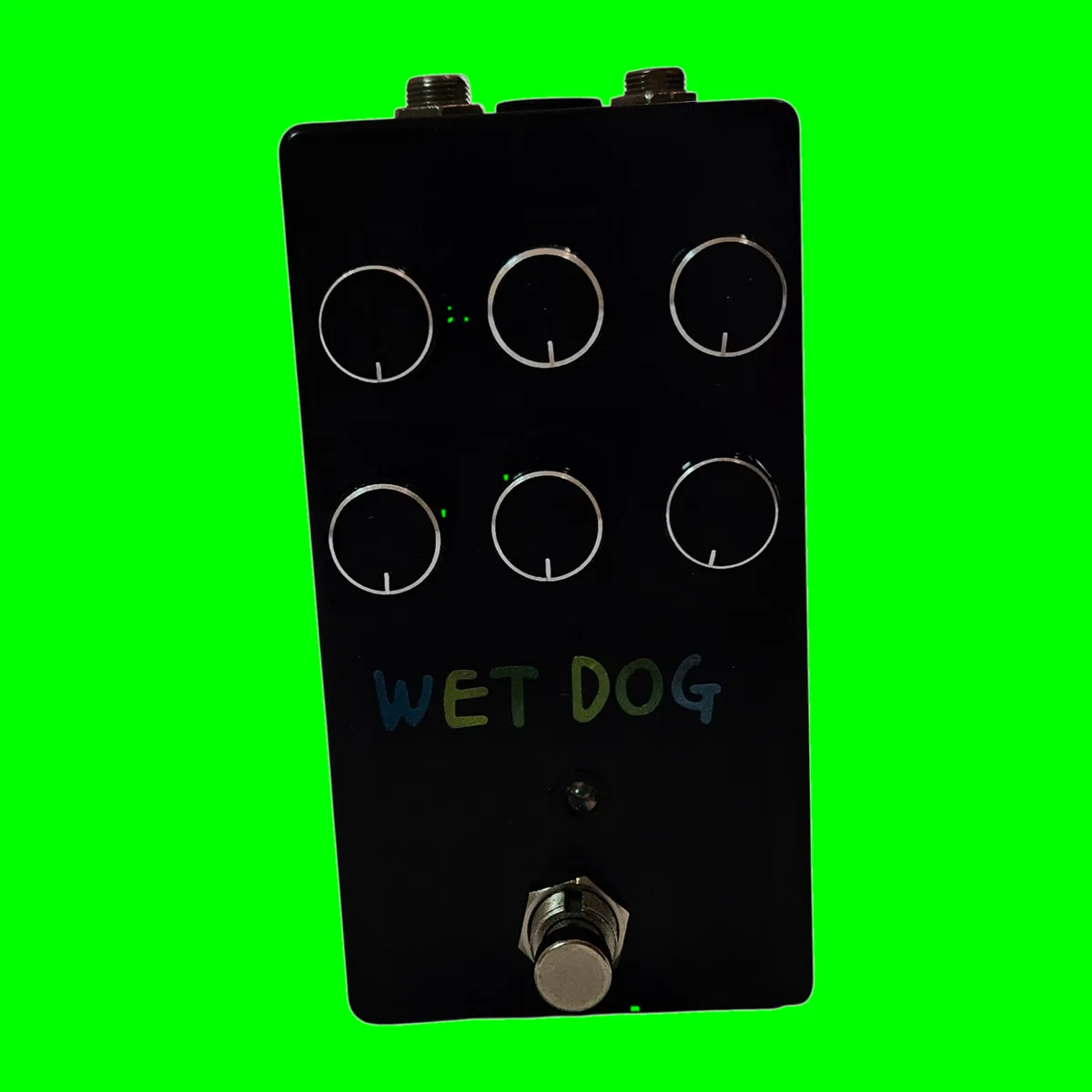 A tall black pedal photographed face on. Six black knobs with thin white pointer lines stand in two rows of three, each ringed by a thin painted circle. WET DOG is hand-lettered below them in blue and green capitals, with a small LED beneath and a silver footswitch at the bottom.
