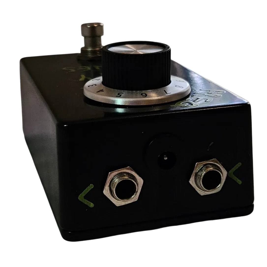 The pedal's top edge straight on, showing two silver quarter-inch jack sockets with a recessed barrel power socket between them. A small hand-painted green arrow beside each jack marks the signal direction.